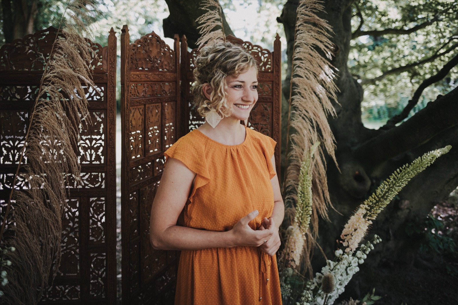 Anne Fraune in einem orangefarbenen Kleid lächelt vor einem geschnitzten Holzparavent und Pampasgras