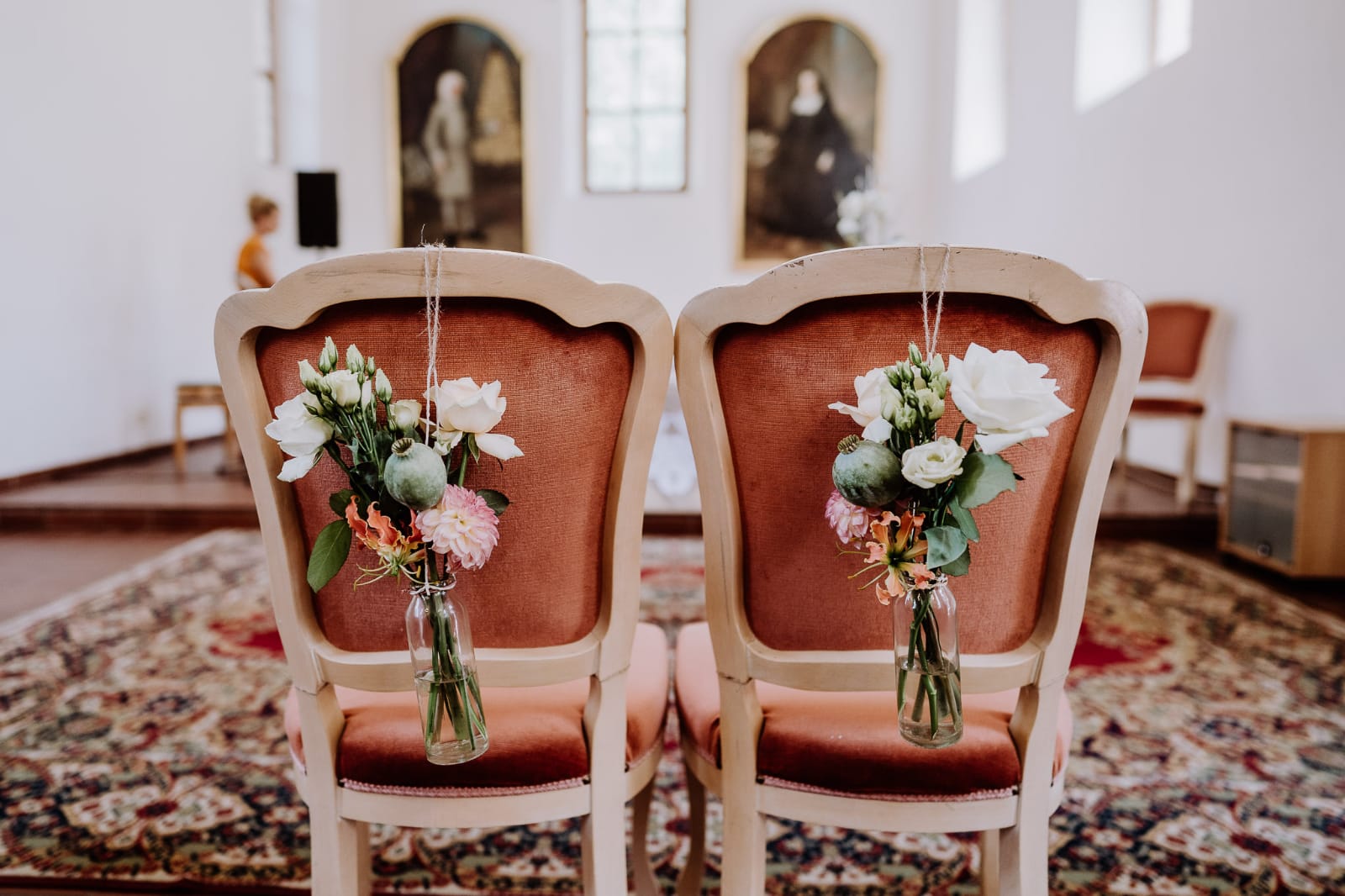 Zwei Stühle für das Brautpaar, geschmückt mit kleinen Blumensträußen in Glasflaschen, in einem Saal mit Teppich und alten Gemälden