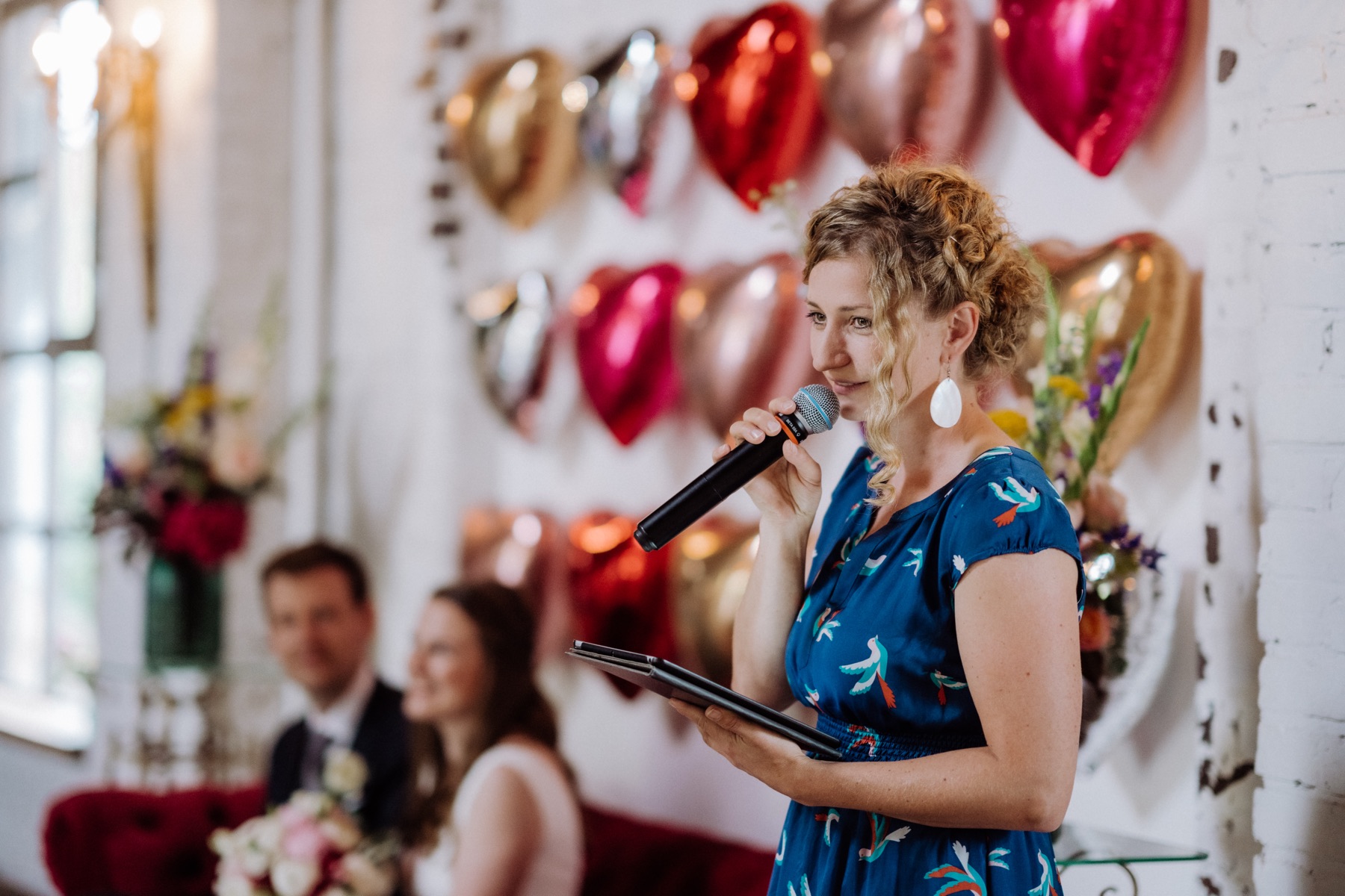 Anne Fraune hält mit Mikrofon und Tablet die Traurede, im Hintergrund sitzt das Brautpaar vor einer Wand aus Herzballons