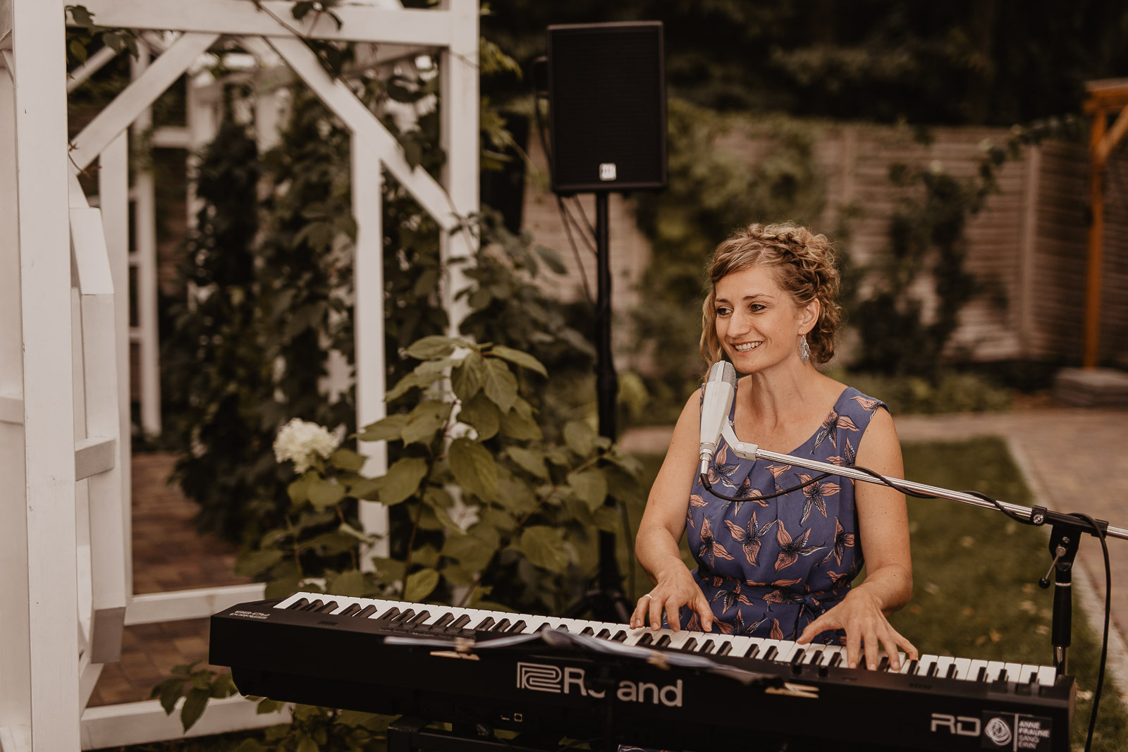 Anne Fraune singt lächelnd am Keyboard im Garten neben einer weißen Pergola