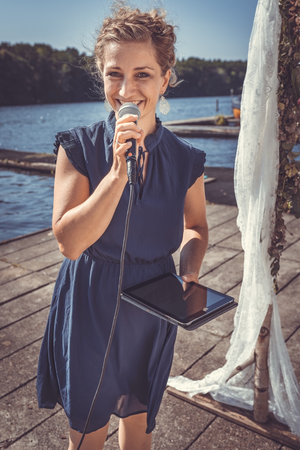 Anne Fraune lächelt auf einem Steg am See, in der einen Hand das Mikrofon, in der anderen ein Tablet, daneben ein Birkenbogen mit weißem Stoff