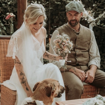 Sarah und Olli sitzen in Korbstühlen, Sarah hält ihren Brautstrauß, davor ihr Hund