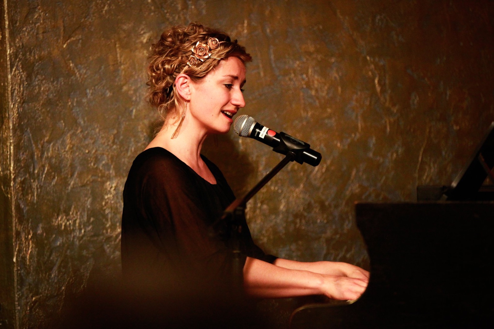 Anne Fraune singt im schwarzen Kleid am Flügel vor einer dunklen, goldschimmernden Wand