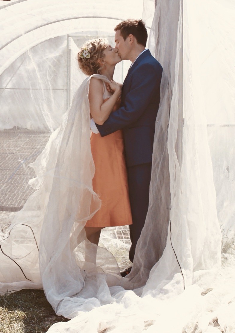 Anne Fraune im orangefarbenen Kleid küsst ihren Mann an ihrem Hochzeitstag, um sie herum wehen weiße Tücher