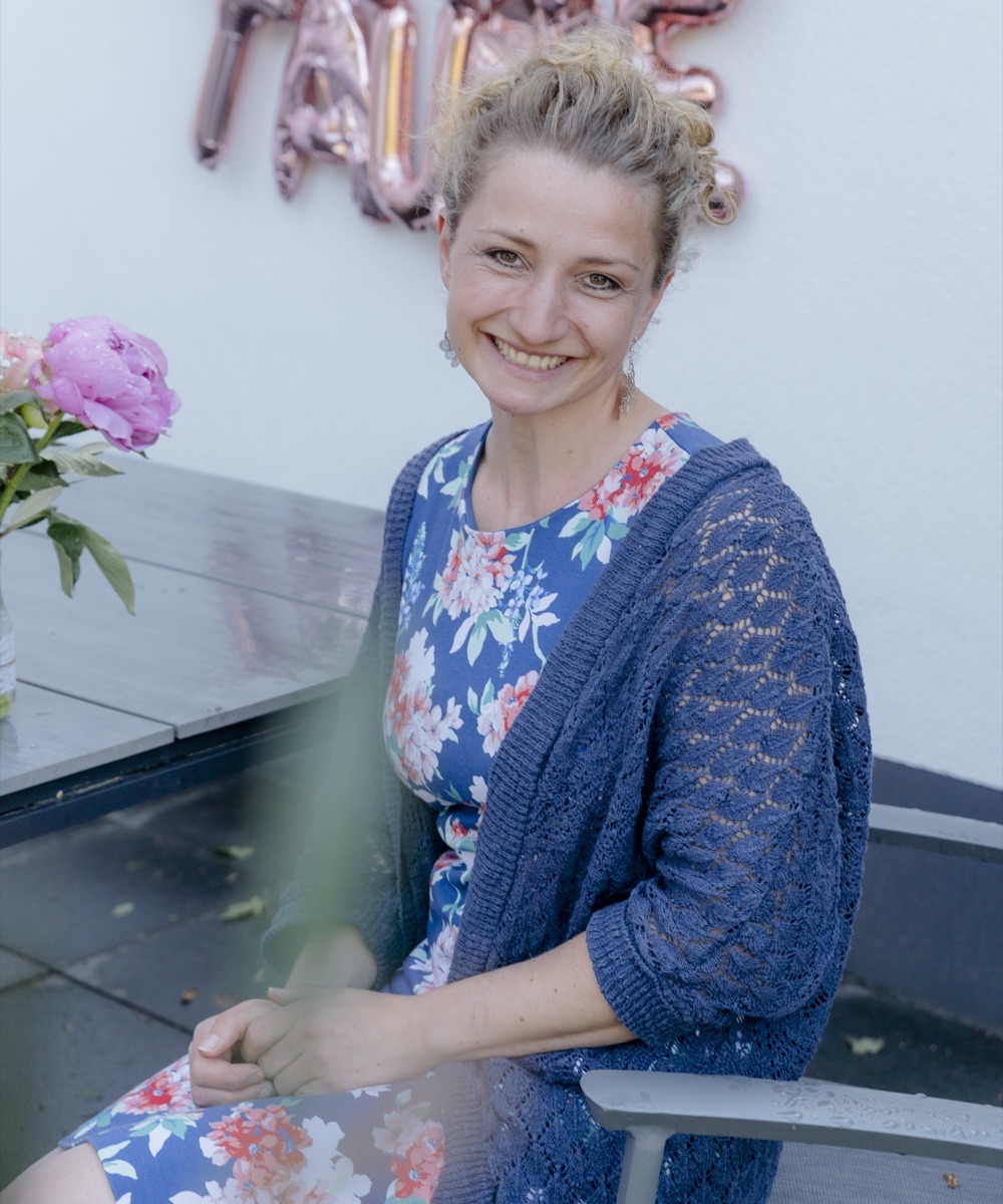 Anne Fraune sitzt lächelnd in einem geblümten Kleid und blauer Strickjacke an einem Tisch mit Pfingstrosen