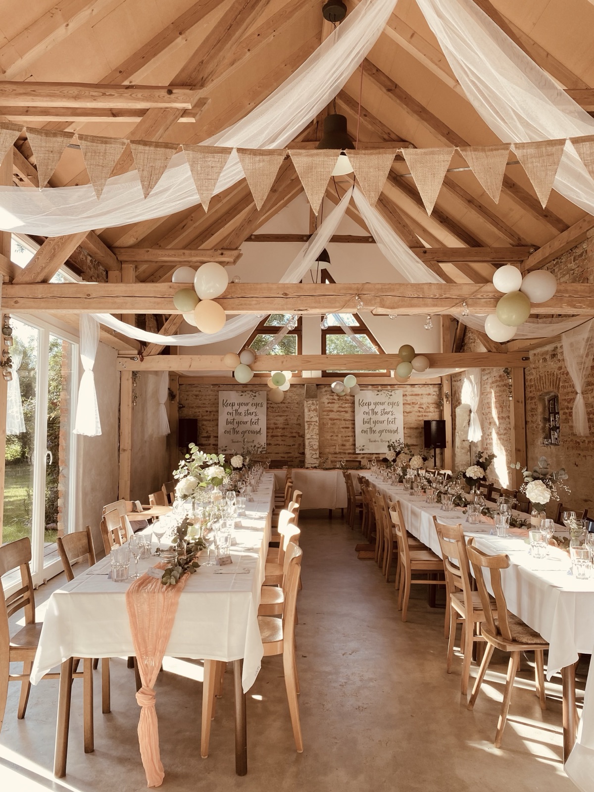 Die Hochzeitsscheune mit langen, weiß gedeckten Tafeln, Wimpelketten und Tüchern unter dem Holzdachstuhl