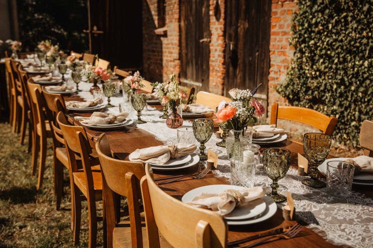Lange gedeckte Holztafel mit Blumen und grünen Gläsern vor der Backsteinwand mit Efeu