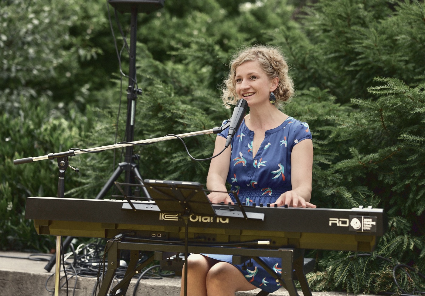Anne Fraune singt lächelnd am Keyboard im Freien vor grünen Bäumen